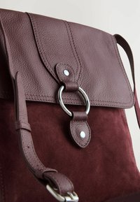 Zaino in pelle e suede bordeaux con superficie testurizzata, chiusura a patta e accentatura a cerchio in metallo. Include una tracolla regolabile.