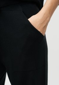 Χέρι που έχει εισαχθεί στην μπροστινή τσέπη μαύρου πλεκτού παντελονιού, δείχνοντας υφή ύφανσης και καρπό με ορατές φλέβες.