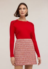 Červený pletený svetr s akcentovanými knoflíky na rameni, zkombinovaný s texturovanou sukní z růžové a červené tweed látky. Jednoduchý design, přiléhavá silueta.