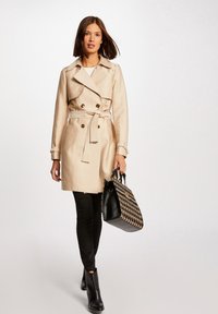 Beige trenchcoat med dubbelknäppt framsida, bälte och axelremmar. Svart mönstrad handväska och figursydda svarta byxor kompletterar looken.