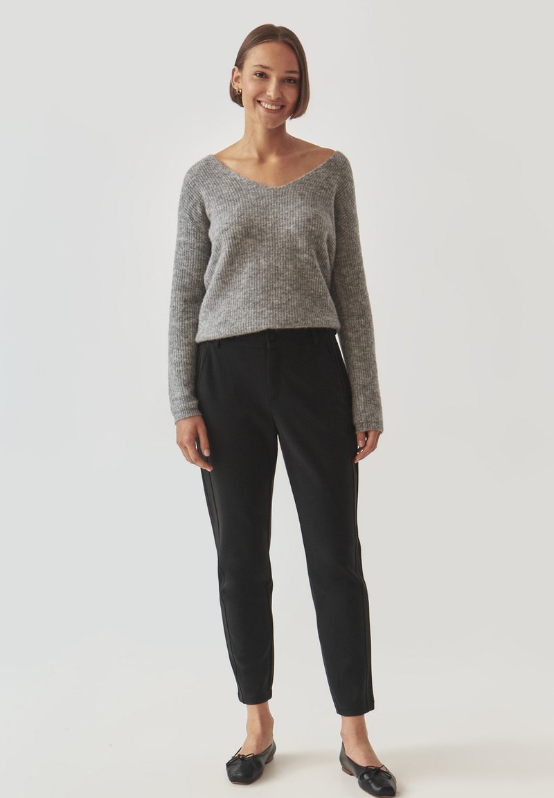 Szary sweter dzianinowy z dekoltem w serek, zestawiony z czarnymi spodniami zwężanymi z bocznymi paskami. Czarne płaskie buty dopełniają zestaw.