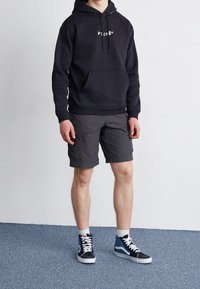 Felpa nera con cappuccio, con tasca frontale e logo bianco, abbinata a pantaloni cargo grigi e scarpe da ginnastica alte blu. Tessuto texturizzato e design casual.