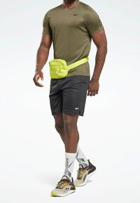 Šedé šortky, olivově zelené tričko a černé běžecké boty se žlutými akcenty. Jasně žlutá crossbody taška je umístěna v pase.