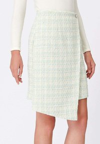 Jupe portefeuille texturée bleu clair et blanche avec un ourlet asymétrique, attachée à la taille par un petit bouton rond, portée par un mannequin.