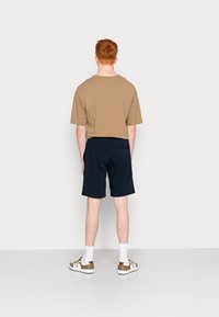 Brown cotton t-shirt with short sleeves, paired with navy shorts. White sneakers with gold accents complete the outfit. Model shown from the back.