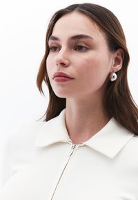 Kremowa, prążkowana bluzka na zamek z kołnierzykiem, z metalowym zamkiem błyskawicznym i gładką, błyszcząca, kroplową bransoletką. Neutralne tło.