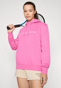 Rosa hettegenser laget av en bomullsblanding med lomme foran; har "BJÖRN BORG" tekstlogo. Lyse shorts med trykket merkevarelogo. Tennisracket i hånden.