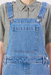 Lys blå denimsal er med justerbare blå stropper og sølvfargete detaljer, og har en frontlomme samt en teksturert grå skjorte under.