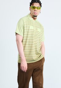 Polo-shirt vert clair avec des rayures horizontales, col côtelé et un logo "AIR" audacieux. Associé à un pantalon marron ample.