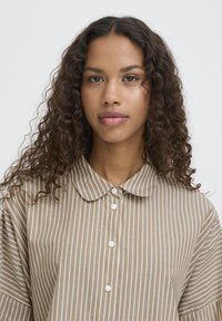 Ung kvinde med langt krøllet hår iført en beige og hvid vertikalt stribet knap-op skjorte med en rund krave.