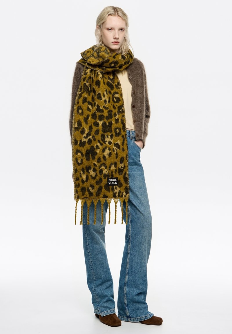 Szalik w panterkę w kolorach oliwkowej zieleni i czerni, z frędzlami na brzegach, noszony na beżowej bluzce i szarym kardiganie, zestawiony z niebieskimi dżinsami.