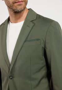 Oliven grøn blazer lavet af tekstureret stof, med knaplukning og en diskret lommetørklæde detaljer. Båret over en hvid t-shirt.