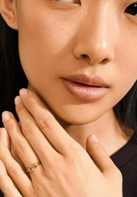 Bague en or avec un design en nœud, portée sur une main reposant contre un visage à la peau lisse. Le fond neutre met en valeur les tons chauds.