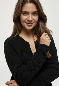 Crna pletena kardigan s teksturiranim uzorkom, okruglim izrezom i rebrastim manžetama, nosi se preko čipkaste bluze, prikazujući bliski prikaz.