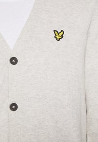 Hellgraue Strickjacke aus weichem Material mit V-Ausschnitt, schwarzen Knöpfen und einem gelben Adler-Logo auf der linken Brust.