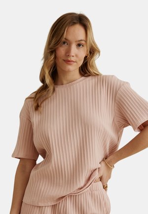 Femme aux longs cheveux blonds portant un haut côtelé rose poudré à manches courtes et un bas assorti, posant avec une main sur la hanche.