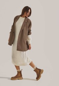 Blazer surdimensionné marron, robe tricotée crème, bottines marron à semelles épaisses. Des textures détaillées et des couleurs contrastantes rehaussent la tenue.