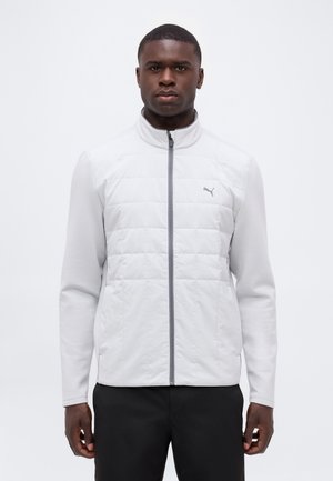 Mann trägt eine weiße gesteppte Jacke mit Reißverschluss und gerippten Ärmeln sowie schwarze Hose, steht vor einem schlichten weißen Hintergrund.