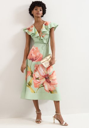 Femme portant une robe midi vert clair avec un grand imprimé floral rose, encolure volantée, tenant une pochette dorée et des sandales à talons beiges.