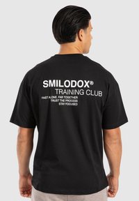 Sort bomulds t-shirt med stor hvid trykt tekst på ryggen, med en afslappet pasform og korte ærmer. Teksten inkluderer mærke og motiverende sætninger.