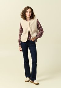 Beige fleeceväst med rundad halsringning och framknäppning, lagd över en blommig långärmad skjorta. Matchas med mörkblå utsvängda jeans och sneakers.