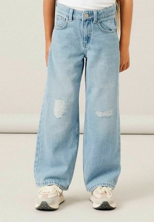 Enfant portant un jean bleu clair à jambes larges avec des déchirures aux genoux et des baskets blanches, debout devant un fond uni.