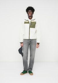 Hvit fleece zip-jakke med grønn brystlapp, grå jeans og grønne sneakers. Outfit inkluderer en svart caps holdt i hånden.