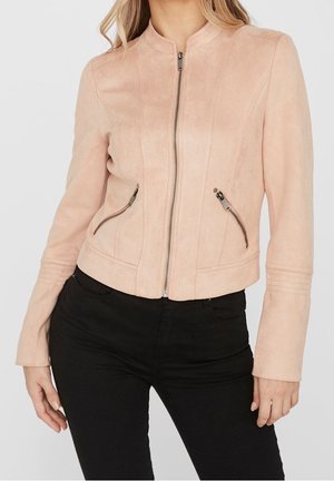 Mujer con chaqueta de ante rosa claro con cremallera y dos bolsillos con cremallera, y pantalones negros, sobre un fondo liso.