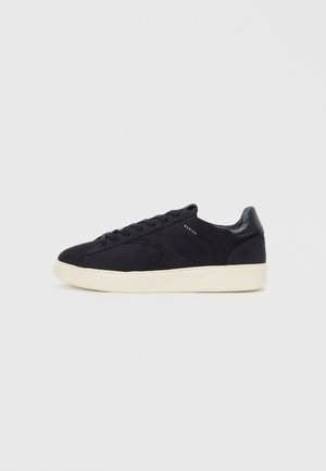 Black suede sneaker with tonal stitching, round toe, flat laces, and a cream rubber sole. Features a glossy black heel accent.
