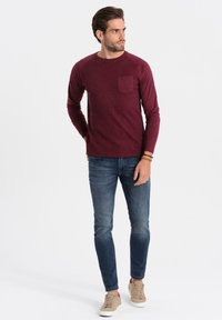 Burgunderfarbendes langärmliges Ripp-Shirt mit einer Brusttasche, kombiniert mit dunkelblauen Jeans und beigen Sneakern. Einfaches, figurbetontes Design.