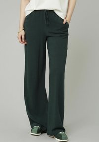 Pantalons larges en tissu vert foncé avec une texture lisse, dotés d'une taille élastique et d'un cordon de serrage, avec des poches latérales.