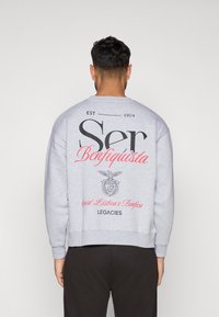 Világosszürke, laza szabású pulóver, nagy fekete felirattal és piros kiegészítőkkel. Tartalmaz egy logót, amelyen egy sas látható és az "Sport Lisboa e Benfica" szöveg.