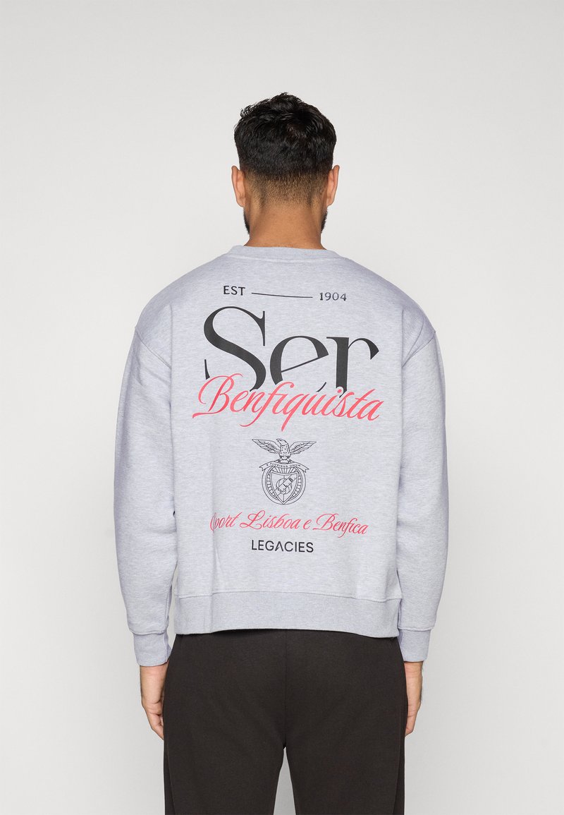 Világosszürke, laza szabású pulóver, nagy fekete felirattal és piros kiegészítőkkel. Tartalmaz egy logót, amelyen egy sas látható és az "Sport Lisboa e Benfica" szöveg.