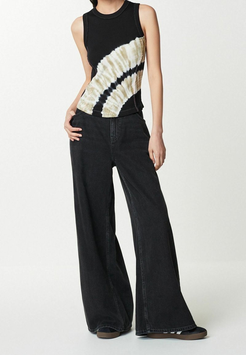 Personne portant un haut noir sans manches avec un motif tie-dye beige et noir et un jean large foncé, debout avec une main dans la poche.