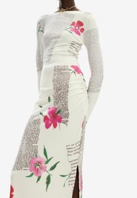 Robe à manches longues en tissu blanc avec des motifs floraux roses et un design de texte de journal ; elle présente des détails froncés sur le côté et une fente sur le côté.