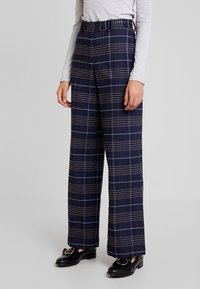 Marineblauwe geruite wijde broek met een bruin en wit ruitpatroon, voorzien van een hoge taille en riemlussen, gecombineerd met zwarte loafers.