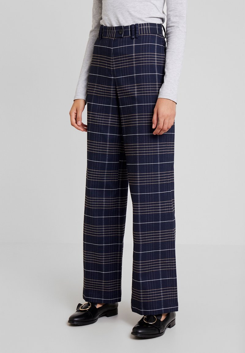 Marineblauwe geruite wijde broek met een bruin en wit ruitpatroon, voorzien van een hoge taille en riemlussen, gecombineerd met zwarte loafers.