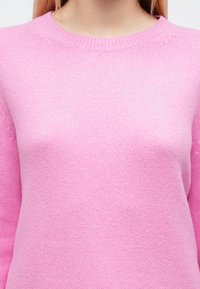 Pulover roz pal tricotat, cu decolteu rotund și margini cu țesătură ribată, având o textură moale și cusături uniforme pe toată suprafața.