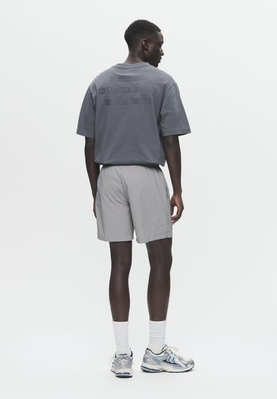 Grå kortærmet t-shirt med tekst på ryggen, sat sammen med lysegrå shorts. Modellen har hvide sokker og sølvfarvede sportsko med blå detaljer på.
