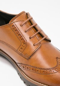 Chaussure oxford en cuir marron avec perforations décoratives, texture lisse, lacets et semelle plate avec une subtile brillance.