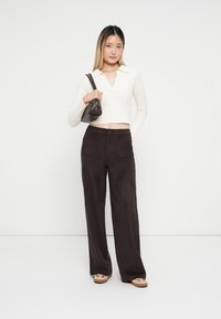 Bijela kabelska pletena kratka majica, tamnosmeđe hlače širokih nogavica, crna torba i svijetle cipele. Glatke teksture i opušten kroj kroz cijeli outfit.