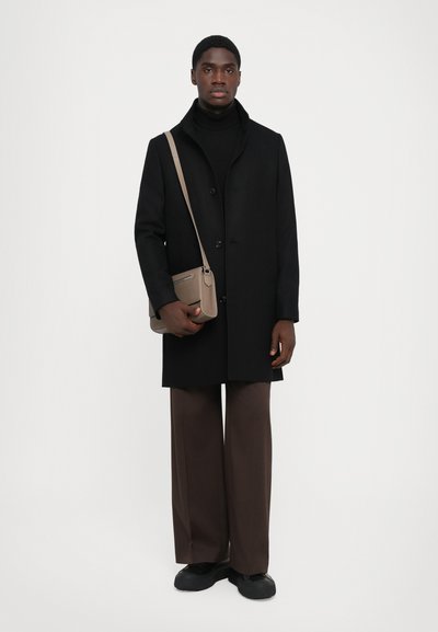 Hombre vestido con abrigo negro, cuello alto negro, pantalones anchos marrones, zapatos negros, y llevando un bolso cruzado beige, contra un fondo blanco.