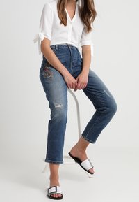 Siniset korkeavyötäröiset farkut kuluneilla yksityiskohdilla, yhdistettynä valkoiseen napilliseen paitaan, jossa on solmittavat hihat, sekä mustavalkoisiin liukusandaaleihin.