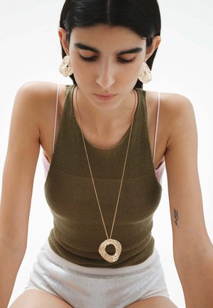 Pendientes de declaración en tono dorado y textura, junto con un collar a juego, adornan a una modelo que lleva una blusa ajustada de color verde oliva y pantalones cortos claros.