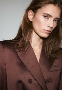 Hnedý saténový blejzer s dvojradovým dizajnom, veľkými gombíkmi a špicatým klopom. Hladká textúra a prispôsobený strih, s jedným vreckom.