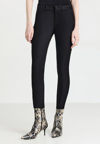 Personne portant un pantalon noir ajusté et des bottines à talons aiguilles en cuir de serpent, se tenant devant un fond uni.
