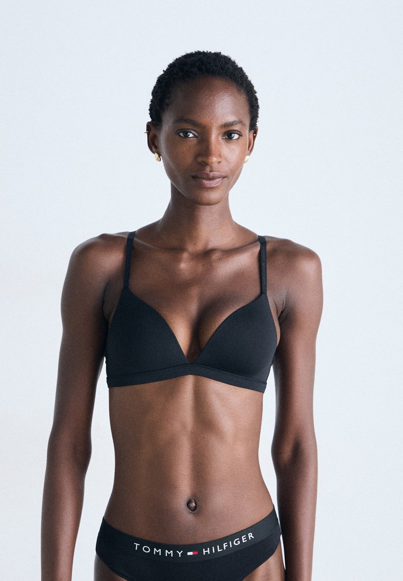 Black triangular bralette with thin straps and smooth texture, paired with black bikini bottoms featuring white and red "TOMMY HILFIGER" text.