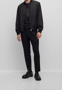 Muž oblečený v černé bombera, černém tričku, černých slim kalhotách a černých kožených šněrovacích botách stojící proti jednoduché šedé zdi.