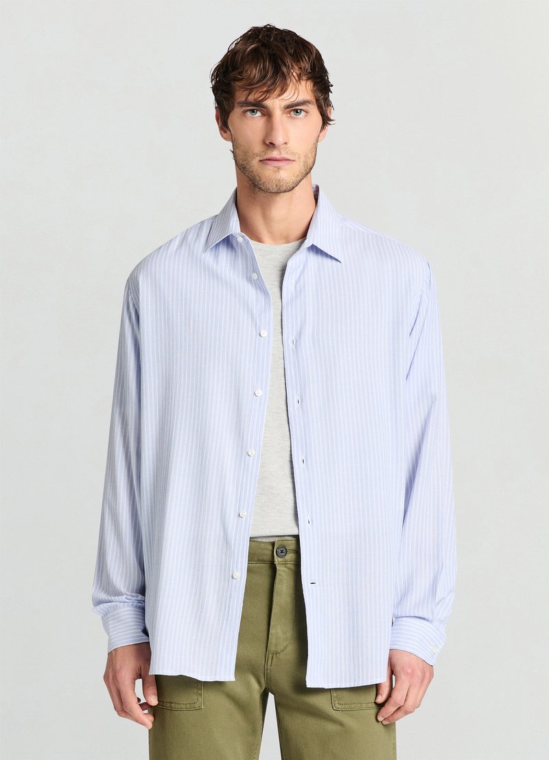 Hellblau gestreiftes Hemd mit Knopfkragen, langen Ärmeln und lockerer Passform. Kombiniert mit einem grauen T-Shirt und olivgrünen Hosen.