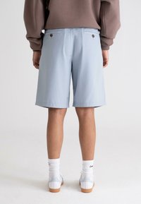 Light blue knee-length shorts with two back pockets featuring brown buttons. Paired with white socks and sneakers on a neutral background.
